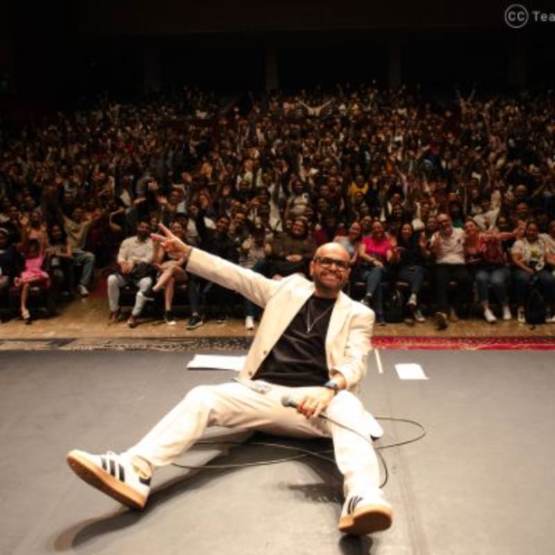 Hombre sentado en el suelo de un escenario posando con el público detrás de él.